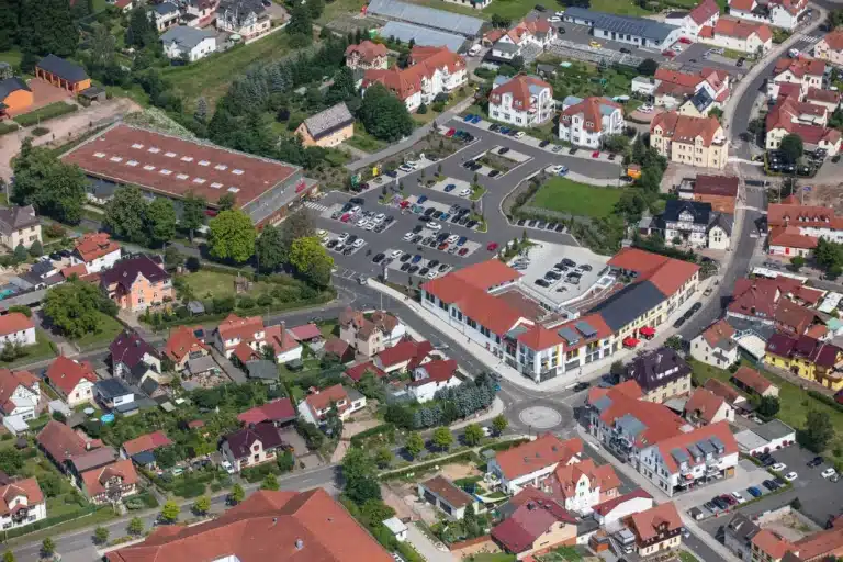 Auf diesem Luftbild ist ein kompletter Stadtkern zu sehen. Dominiert wird dieser durch rote Dacheindeckungen. Die Bebauung besteht überwiegend aus Ein- und Mehrfamilienhäusern mit Gärten, Hecken und Bäume. In der Mitte des Bildes befindet sich ein Straßenkreisel und direkt dahinter ein sehr großes Wohn- und Geschäftshaus und einem danebenliegenden Lebensmittelmarkt. Zwischen diesen Gebäude befindet sich eine große Parkfläche mit Bäumen und Grünanlage. Durch den Stadtkern verlaufen einige Straßen und Wege.