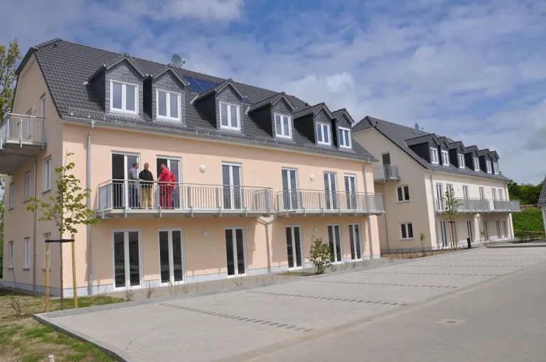 Auf dem Foto sind zwei baugleiche Wohngebäude zu erkennen. Die Gebäude bestehen aus Erdgeschoss, 1. Obergeschoss und ausgebautem Dachgeschoss. Im Erdgeschoss befinden sich Terrassen, im Ober- und Dachgeschoss (Giebel) großzügige Balkone. Jedes Haus wurde mit jeweils sechs Gauben und weißen Fenstern ausgestattet. Das Erd- und Obergeschoss verfügen über bodentiefe weiße Fenster. Das Dach ist mit grauen Ziegelsteinen eingedeckt. Das linke Haus hat einen Anstich in apricot und das rechte in zartem gelb. Die Flächen vor den Häusern sind gepflastert und als PKW Stellplätze abgebildet. Auf einem Balkon befinden sich vier Personen. Vor jedem Haus wurde ein Baum gepflanzt. Der Himmel ist blau mit einigen Wolken.