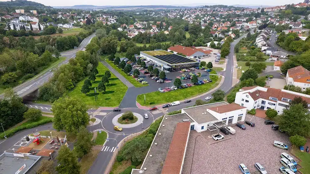 Das Bild zeigt eine Luftaufnahme eines städtischen Gebiets mit einem Supermarkt in der Bildmitte, dessen Dach teilweise mit einer Solaranlage bedeckt ist. Vor dem Gebäude befinden sich zahlreiche Parkplätze. Ein Kreisverkehr mit mehreren einmündenden Straßen liegt im Vordergrund. Um den Supermarkt herum erstreckt sich ein grünes Gebiet mit Bäumen. Im Hintergrund sind zahlreiche Wohnhäuser und eine hügelige Landschaft zu sehen. Unten rechts befindet sich ein Gebäudekomplex mit mehreren Polizei- oder Einsatzfahrzeugen. Links unten ist ein kleiner Spielplatz mit rotem Spielgerät erkennbar.