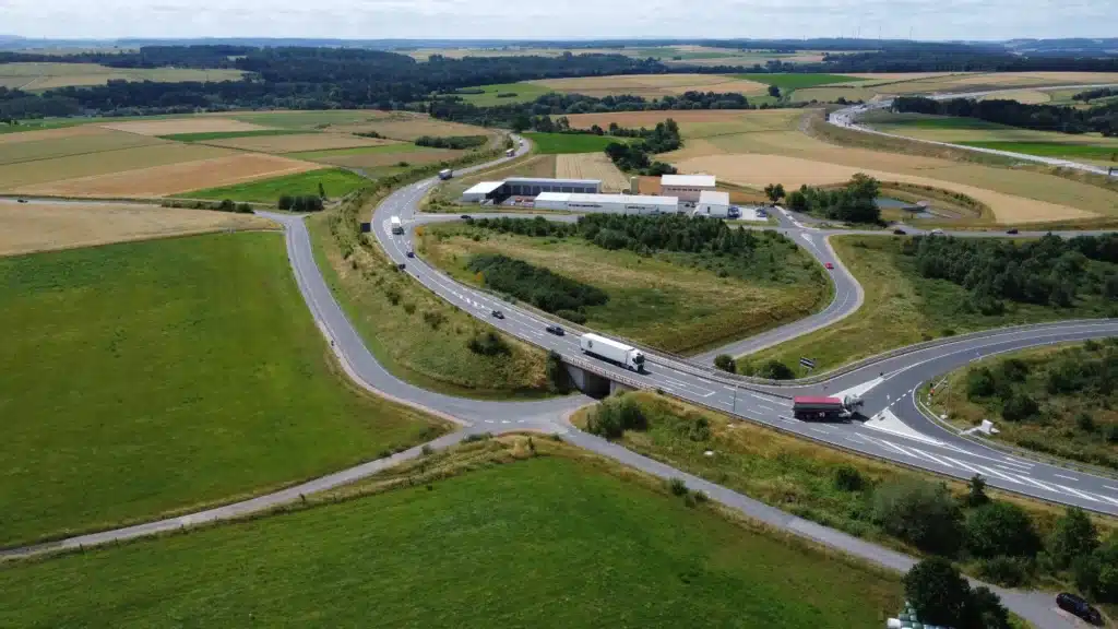 Auf dieser Luftaufnahme ist eine grüne Landschaft mit Feldern, Wiesen, Wäldern und einigen sich kreuzenden Straßen zu sehen. Ebenfalls ist mittig im Hintergrund der Straßen ein Hallenensemble zu erkennen. Auf den Fahrbahnen sind einige LKW's und PKW's unterwegs. Am rechten hinteren Bildrand erkennt man schemenhaft eine Autobahn.
