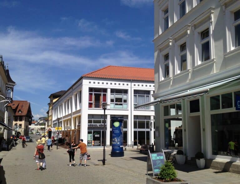 Auf dieser Visualisierung ist eine verkehrsberuhigte gepflasterte Geschäftsstraße zu erkennen. Rechts und links der Straße befinden sich Geschäftshäuser mit großen Schaufenstern, Markisen, Vordächern, Werbeanlagen und teilweise mit Blumenkübeln. Durch die Straße laufen Menschen in sommerlicher Kleidung. Der Himmel ist blau. Im Vordergrund erkennt am eine Straßenlaterne und eine Litfaßsäule.