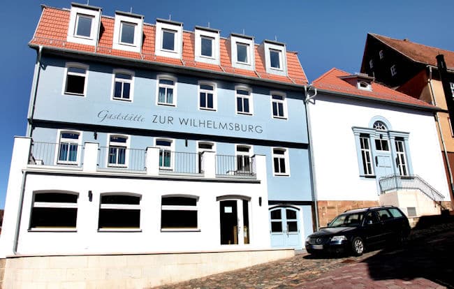 Auf dem Bild ist ein viergeschossiges Gebäude mit angrenzendem Nebengebäude zu sehen. Das Gebäude ist komplett saniert. Die Fassade ist hellblau gestrichen die Sprossenfenster sind weiß. Im Erdgeschoss gibt es einen in weiß gestrichenen Vorbau von circa 5 Metern Breite mit drei großen Fenstern und einer Tür mit Glaselement. Die darüberliegende Fläche dient als Balkon für das erste Obergeschoss. Rechts neben dem Vorbau befindet sich im Erdgeschoss eine doppelflügelige ebenfalls in blau gestrichene Eingangstür mit Glas. Im Dach wurden sechs Gauben eingebaut. Das direkt angrenzende Nebengebäude mit weißer Fassade und rotem Ziegeldach verfügt über eine Außentreppe mit Sprossengeländer sowie einer in blau gestrichenen Eingangstür mit direkt angrenzenden Sprossenfenstern rechts und links von der Tür und einem halbrunden Oberlicht direkt über der Tür. Im Dach befindet sich eine sehr kleine Gaube. Vor dem Haus ist eine gepflasterte Straße und Gehweg mit starkem Gefälle erkennbar, sowie ein dunkler PKW.