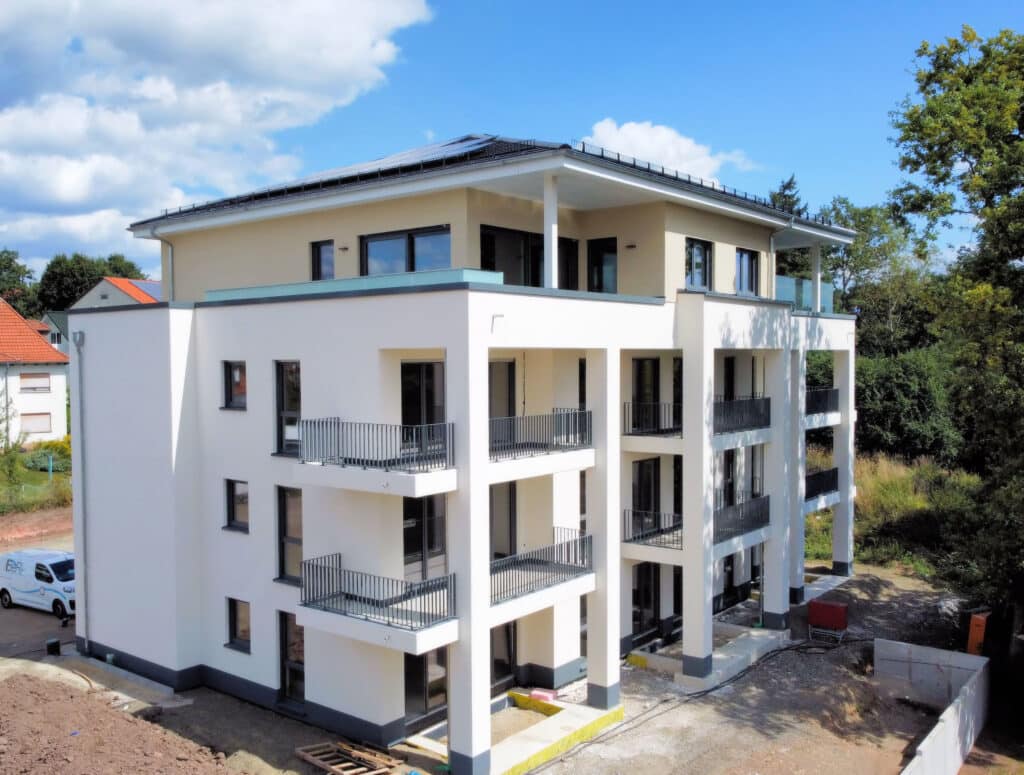 Das Bild zeigt ein modernes Wohnhaus mit insgesamt 11 Wohneinheiten. Jede Wohnung verfügt über einen großzügigen Balkon. Das Erdgeschoss sowie das 1. und 2. Obergeschoss sind identisch. Hier befinden sich pro Etage drei Wohnungen. Das Obergeschoss besteht aus zwei Penthouse-Wohnungen mit Dachterrassen. Die Außenanlage befindet sich noch in der Fertigstellung.