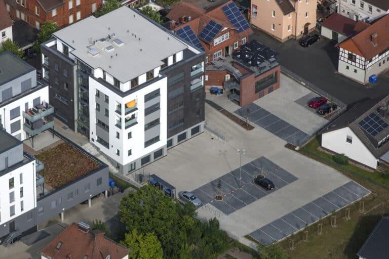 Auf dieser Drohnenaufnahme ist ein fünfgeschossiges Gebäude mit Flachdach zu sehen. Das moderne Gebäude ist mit Balkonen und Dachterrassen ausgestattet. Die Fassade ist in weiß, grau und anthrazit gestrichen. Die Fenster sind ebenfalls anthrazitfarben. Im Untergeschoss sind fünf Garagen eingebaut. Vor den Garageneinfahrten befindet sich eine gepflasterte Parkplatzanlage für 24 PKW. Die Nachbarbebauung ist sehr eng zu unserem Gebäude. Die Aufnahme zeigt die Hinteransicht der Gebäude mit Gärten. Die Gebäude rechts im Bild sind teilweise Wohnhäuser oder Mehrfamilienhäuser mit Fachwerk, Putz- beziehungsweise Klinkerfassaden und asphaltierten Flächen rund um die Häuser. Links im Bild ist ebenfalls ein modernes mehrgeschossiges Gebäude an Anstellbalkonen und Flachdach zu sehen. Im Bild sind vereinzelt Bäume erkennbar.