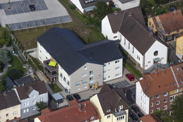 Auf dieser Drohnenaufnahme ist eine enge Wohnbebauung zu sehen. In der Mitte dominiert ein großes Wohngebäude mit Anstellbalkon, schwarzer Dacheindeckung ohne Dachfenster und einer hellen Fassade. Der Eingang des Gebäudes ist mit einem Glasvordach überbaut und dieser Bereich ist orangefarben gestrichen. Die Fenster sind in weiß ausgeführt. Bei einigen Fenstern sind geschlossene Rollos zu sehen. Im hinteren Bild ist eine Parkplatzanlage mit Grünstreifen und Baumreihe zu sehen. Rechts neben dem Gebäude ist eine Kirche mit Wohnhausanbau zu erkennen, sowie zwei Gebäudeteile in gelb teilweise mit Fachwerk. Im Vordergrund sind mehrgeschossige Wohngebäude älteren Baujahrs zu sehen. Die Dächer sind teileweise mit roten oder dunklen Dachziegel eingedeckt. Ebenfalls sind Teile einer Straße erkennbar. Im linken Bild sind direkt an der Straße verlaufende Häuser mit dahinterliegenden Gärten zu erkennen.