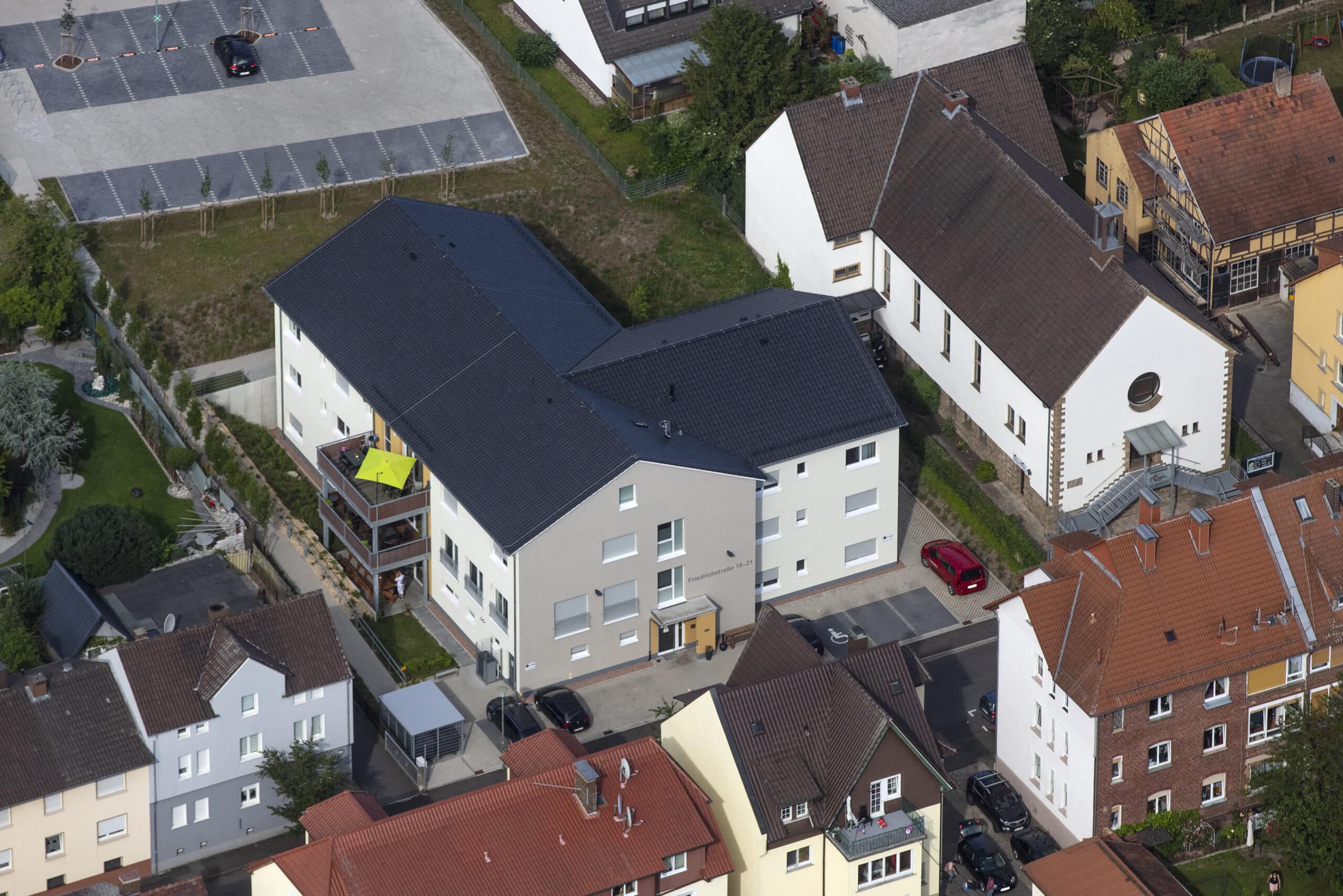 Auf dieser Drohnenaufnahme ist eine enge Wohnbebauung zu sehen. In der Mitte dominiert ein großes Wohngebäude mit Anstellbalkon, schwarzer Dacheindeckung ohne Dachfenster und einer hellen Fassade. Der Eingang des Gebäudes ist mit einem Glasvordach überbaut und dieser Bereich ist orangefarben gestrichen. Die Fenster sind in weiß ausgeführt. Bei einigen Fenstern sind geschlossene Rollos zu sehen. Im hinteren Bild ist eine Parkplatzanlage mit Grünstreifen und Baumreihe zu sehen. Rechts neben dem Gebäude ist eine Kirche mit Wohnhausanbau zu erkennen, sowie zwei Gebäudeteile in gelb teilweise mit Fachwerk. Im Vordergrund sind mehrgeschossige Wohngebäude älteren Baujahrs zu sehen. Die Dächer sind teileweise mit roten oder dunklen Dachziegel eingedeckt. Ebenfalls sind Teile einer Straße erkennbar. Im linken Bild sind direkt an der Straße verlaufende Häuser mit dahinterliegenden Gärten zu erkennen.