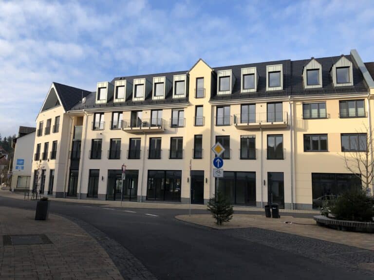 Auf dem Foto erkennt man ein 4-geschossiges Gebäude welches im Erdgeschoss und 1. Obergeschoss gewerblich genutzt wird. Das 2. Obergeschoss, sowie das Dachgeschoss mit Gauben ist als Wohnraum ausgebaut. Zwei Balkone im 2. Obergeschoss sind zu sehen. Das Gebäude ist symmetrisch mit großen Fensterflächen auf jeder Etage versehen. Die Fassade wurde in zarten beige/gelb Tönen gestrichen. Im Vordergrund des Gebäudes erkennt man die Bahnhofstraße mit Gehsteig und Parkbucht. Ebenfalls im rechten Bildrand ist ein Baum eingegrenzt mit einer umliegenden Sitzbank. Im Vordergrund des Bildes ist ein Straßenschild zu sehen. An diesem Schild ist ein Tannenbaum befestigt. Der Himmel ist blau mit zarten weißen Wölkchen.