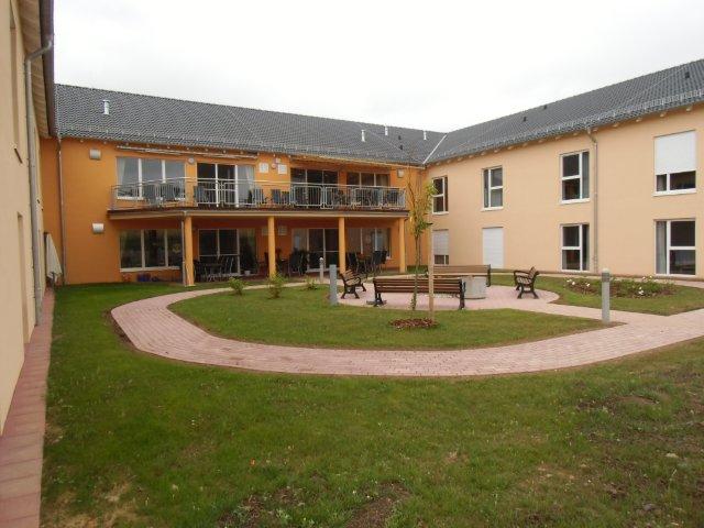 Auf dem Bild ist ein zweigeschossiges Gebäude in U-Form zu sehen. Die Fassade der Seitengebäude rechts und links ist in beige der Mittelteil in einem orange gestrichen. Der mittlere Gebäudeteil hat einen großen Balkon, welcher mit vier Säulen gestützt wird. Das Balkongeländer ist als Stabgeländer ausgeführt. Auf dem Balkon befinden sich circa 16 Stühle und mehrere Tische. Der Innenhof ist großflächig mit Rasen ausgestattet. In der Mitte der Grünanlage befindet sich eine circa 25 m²rund gepflasterte Fläche mit vier Sitzbänken. Rechts neben der Sitzfläche ist ein Blumenbeet angelegt, links drei Büsche und im Vordergrund ein junger Baum. Um das Rondell in der Mitte führt ein gepflasterter Fußweg.