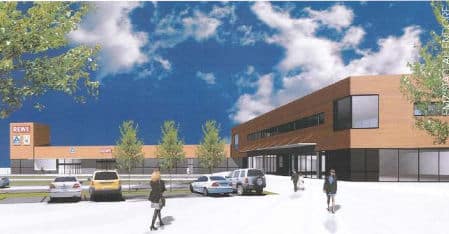 Auf dem Bild ist ein modernes zweigeschossiges Geschäftsgebäude mit Flachdach zu erkennen. Im Erdgeschoss befinden sich große Schaufensterfronten und im Obergeschoss Fensterbänder. Vor dem Gebäude in L-Form befinden sich Parkplätze, einige Bäume, PKW's und Fußgänger. Die Fassaden sind mit Trespa-Platten im oberen Bereich mit Eiche hell und im unteren Bereich in anthrazit verkleidet. Am linken Gebäudeende befindet sich ein Turm mit Werbeaufschriften Rewe, Aldi und Müller.