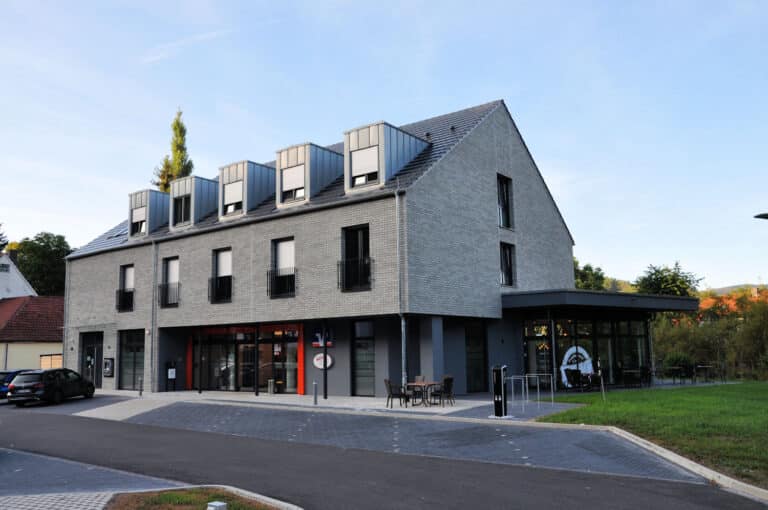 Auf diesem Bild ist ein moderner zweigeschossiger Baukörper mit ausgebautem Dachgeschoss und fünf Gauben zu sehen. Das Gebäude und die Fenster sind komplett in Grautönen gehalten. Die Fassade ist mit grauen Klinkern versehen. Es gibt keinen Dachüberstand. Im Untergeschoss befindet sich ein Cafè mit bodentiefen Fenstern sowie eine Glas-Eingangstür. Diese ist mit zwei roten schmalen Paneelen rechts und links neben der Tür versehen. An der rechten Giebelseite des Gebäude wurde im Erdgeschoss ein kleiner Wintergarten angebaut. Links am Gebäude befindet sich ein weiterer Eingang mit Glastür und kleinem Glasvordach. Im Obergeschoss befinden sich fünf bodentiefe Fenster mit Französischem Balkon. An der Giebelseite befinden sich im 1. Obergeschoss und im Dachgeschoss jeweils ein Fenster. Rechts neben dem Gebäude ist eine kleine Grünfläche, im Hintergrund der Grünfläche und dem Gebäude erkennt am Bäume und ein rotes Dach mit Schornstein. Vor dem Gebäude befinden sich sieben Parkplätze sowie eine kleine Zufahrtstraße.