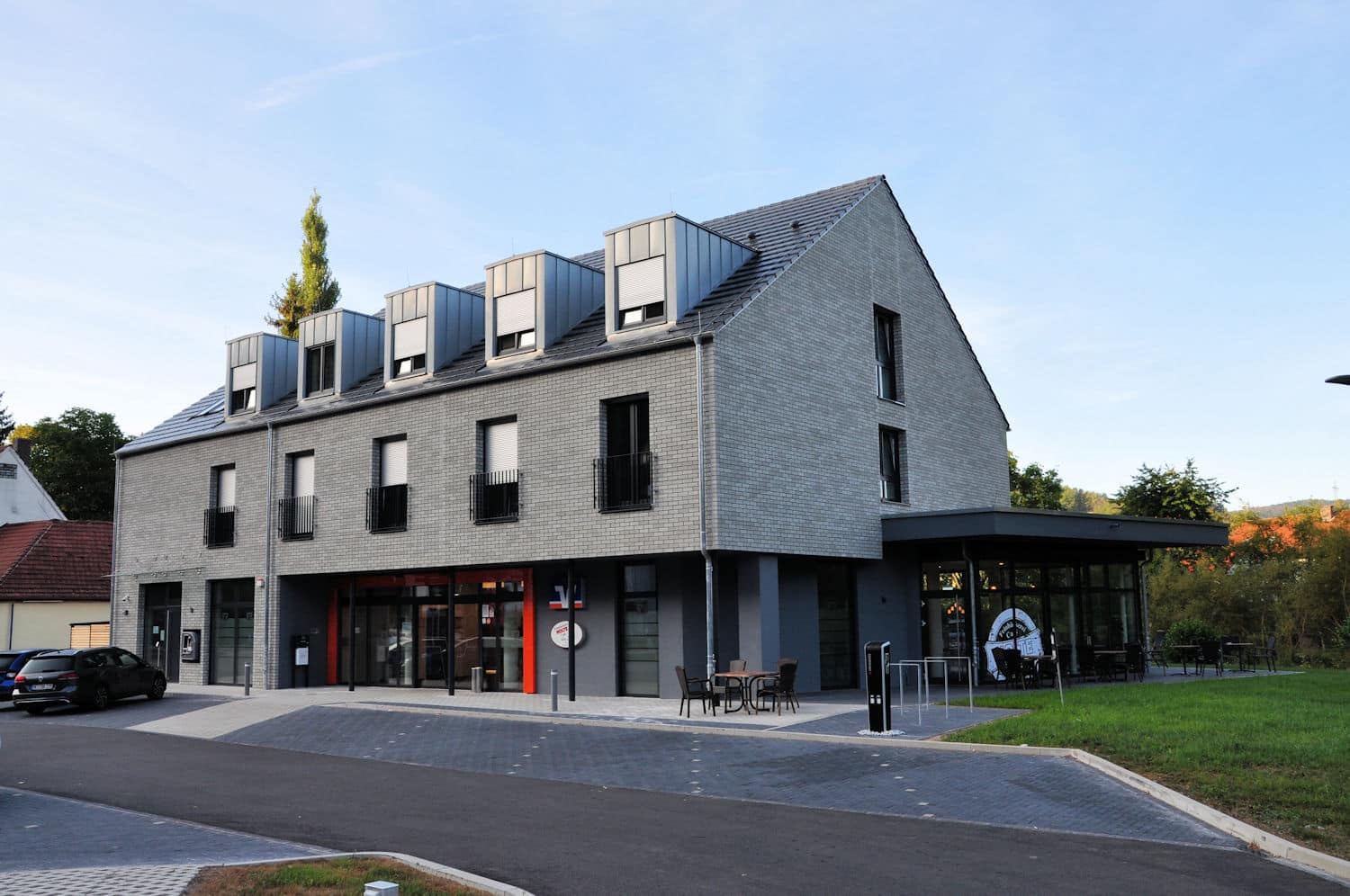 Auf diesem Bild ist ein moderner zweigeschossiger Baukörper mit ausgebautem Dachgeschoss und fünf Gauben zu sehen. Das Gebäude und die Fenster sind komplett in Grautönen gehalten. Die Fassade ist mit grauen Klinkern versehen. Es gibt keinen Dachüberstand. Im Untergeschoss befindet sich ein Cafè mit bodentiefen Fenstern sowie eine Glas-Eingangstür. Diese ist mit zwei roten schmalen Paneelen rechts und links neben der Tür versehen. An der rechten Giebelseite des Gebäude wurde im Erdgeschoss ein kleiner Wintergarten angebaut. Links am Gebäude befindet sich ein weiterer Eingang mit Glastür und kleinem Glasvordach. Im Obergeschoss befinden sich fünf bodentiefe Fenster mit Französischem Balkon. An der Giebelseite befinden sich im 1. Obergeschoss und im Dachgeschoss jeweils ein Fenster. Rechts neben dem Gebäude ist eine kleine Grünfläche, im Hintergrund der Grünfläche und dem Gebäude erkennt am Bäume und ein rotes Dach mit Schornstein. Vor dem Gebäude befinden sich sieben Parkplätze sowie eine kleine Zufahrtstraße.