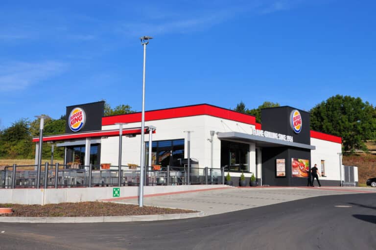 Auf dem Bild ist ein neues modernes Fast Food Restaurant von Burger King zu sehen. Das Gebäude ist weiß gestrichen und am oberen Rand zum Flachdach rot abgesetzt. Drei große Fenster dominieren das Gebäude. Der Eingangsbereich auf der Längsseite des Gebäudes ist überdacht und rechts mit einem anthrazitfarbenen Vorbau versehen. Auf diesem Vorbau ist das Logo von Burger King angebracht sowie im unteren Bereich zwei Werbeschilder. Links vom Eingang sind drei kleine Nadelbäume gepflanzt. Auf der Stirnseite des Gebäudes befindet sich eine große mit Glas umzäunte Terrasse. Diese kann durch eine in der Mitte angebrachte rote Markise beschattet werden. Ebenfalls an dieser Stirnseite befindet sich ein weiteres Logo Burger King auf anthrazitfarbenem Hintergrund. Im vorderen Bereich des Bildes ist die Zufahrt zu den gepflasterten Parkplätzen zu erkennen. Im Kreuzungsbereich Burger King zur Straße befindet sich auf einer kleinen Erdfläche eine Straßenlampe. Eine Person verlässt gerade den Eingangsbereich Richtung Parkplatz. Der Himmel ist strahlend blau. Im Hintergrund sind große Bäume zu erkennen.
