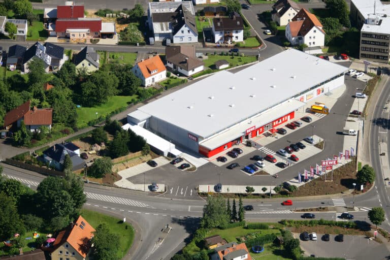 Das Luftbild zeigt in der Mitte des Bildes einen modernen Rewe Markt mit Parkplatzanlage. Im Vordergrund sind zwei Kreuzungen mit Zebrastreifen und Abbiegerspuren zu erkennen, sowie zwei Wohnhäuser mit Grünanlagen. Im rechten hintern Bild erkennt man das mehrgeschossige Rathaus. Im Hintergrund des Rewe Marktes befinden sich weitere Wohnhäuser und Straßen mit Grünanlagen.