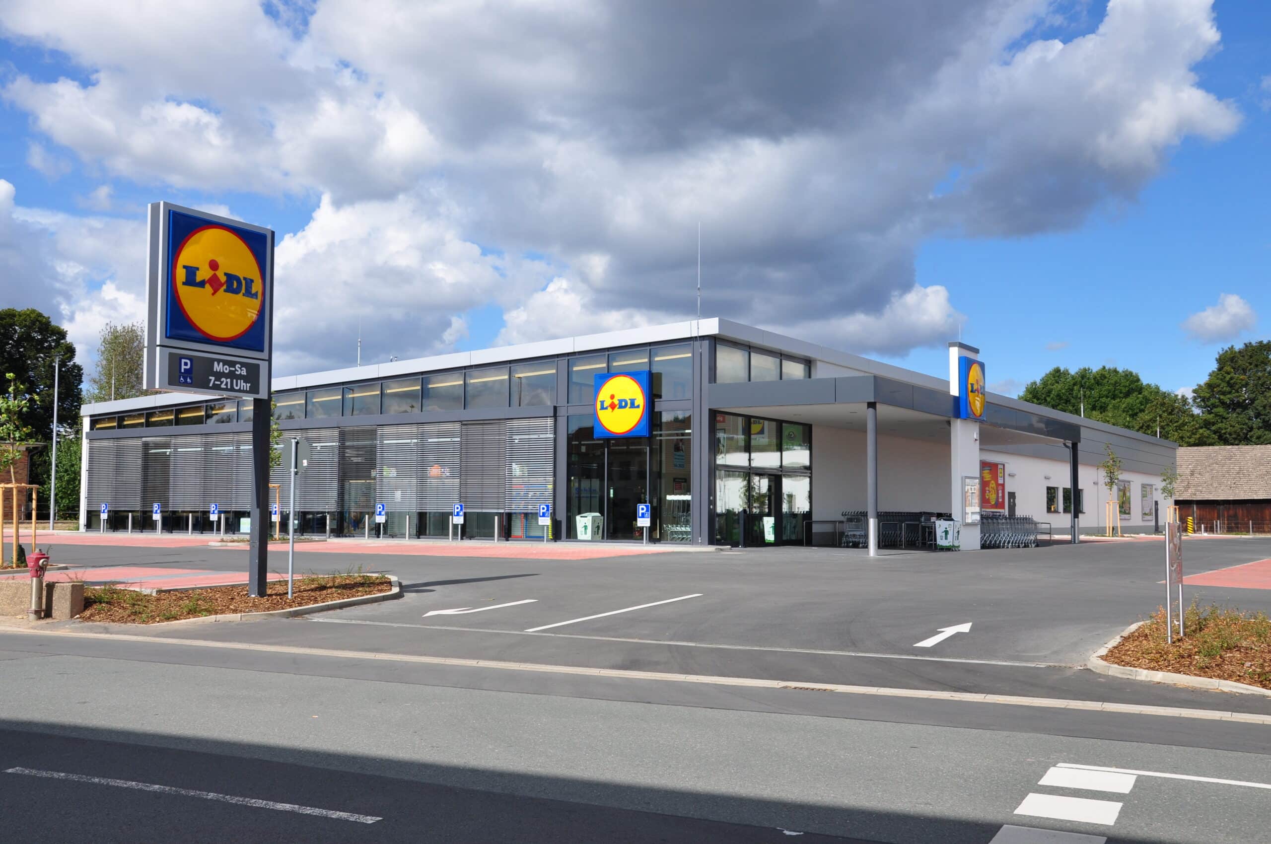 Auf diesem Bild sieht man einen Lidl Lebensmittelmarkt. Der neue und moderne Markt ist hat ein Flachdach und eine komplette Glasfront an der Stirnseite. Der Eck-Eingangsbereich ist überdacht ebenso die Einkaufswagen. Die verputzen Wände des Marktes sind weiß der obere Teil des Gebäudes und die Fensterrahmen sind in grau ausgeführt. An der Glasfront ist eine Beschattung angebracht, teilweise sind hier die Jalousien heruntergefahren. Vor dieser Glasfront befinden sich Parkplätze für behinderte Menschen welche rot gepflastert sind. Die Fahrbahnen vor und neben dem Markt auf dem Grundstück sind asphaltiert und mit Fahrtrichtungspfeilen versehen. Über dem Eingang und an dem Vordach sind große Lidl Werbelogos angebracht. Im Vordergrund des Bildes an der Einfahrt zum Markt befindet sich ein Werbepylon mit Logo und Öffnungszeiten.