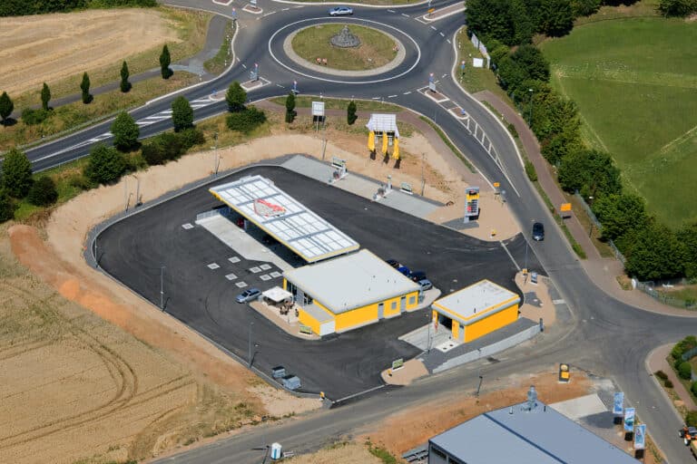 Auf dem Luftbild ist eine Großtankstelle zu sehen. Das Gewerbegrundstück ist umschlossen von Straßen. Die Tankstelle steht auf einem großen asphaltiertem Grundstück und besteht aus einem offenen überdachten Tankbereich, einem großen Nebengebäude mit Terrasse und einer separaten Waschanlage. Die Fassaden der Gebäude sind gelb gestrichen. Rechts von der Waschanlage ist die Einfahrt auf das Grundstück, links die Ausfahrt. Die Fassaden sind in gelb gestrichen. Rechts hinten befinden sich acht PKW-Stellplätze auf dem Anwesen. Die Fläche von diesen Parkplätzen zum Straßenrand ist noch nicht begrünt allerdings sind dort bereits Fahnenmaste mit gelben Fahnen aufgestellt. Im Hintergrund der Tankstelle ist ein sehr großer Verkehrskreisel zu sehen. Hier gehen vier Straßen ab. Die Straßen sind mit Markierungen und Straßenschildern versehen. Die Nachbargrundstücke sind nicht bebaut hier sieht man Äcker und Wiesen. Nur das gegenüberliegende Grundstück ist scheinbar mit einer größeren Halle bebaut. Rechts neben diesem Gebäude befinden sich drei Fahnenmaste mit Werbeaufdruck. Die Straßen zum Kreisel sind teilweise mit Bäumen und Hecken bepflanzt.