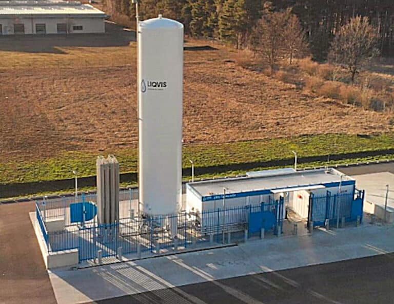 Auf dieser Drohnenaufnahme erkennt man eine Tankstelle für alternativ Kraftstoffe. Die Tankstelle steht auf einem Planum und ist eingezäumt mit einem blauen Doppelstabmattenzaun. Im Inneren befindet sich ein rechteckiger Container mit Durchgang. Links neben dem Gebäude befindet sich ein aufrechter circa 10 Meter hoher Gastank. Im Hintergrund ist eine abgemähte Wiese zu sehen.