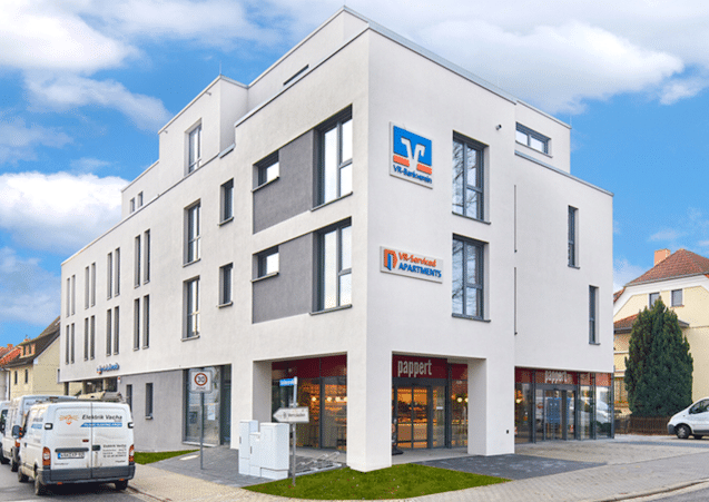 Auf diesem Foto ist ein moderner Baukörper mit vier Geschossen und Flachdach abgebildet. Im Erdgeschoss befindet sich an der Frontseite zum gepflasterten Gehweg etwas zurück gebaut eine Filiale der Bäckerei Pappert sowie im hinteren Bereich des Gebäudes eine Geschäftsstelle der VR-Bank. Das Gebäude ist weiß gestrichen mit grauen Akzenten im ersten und zweiten Obergeschoss. Das Erdgeschoss ist im Bereich Bäckerei über die gesamte Breite Richtung Straße mit großen Fensterflächen ausgestattet. Vor diesen großen Fensterflächen befinden sich mehrere Parkplätze, eine sehr kleine Grünfläche sowie drei Verteilerkästen. Der links zu sehende Erdgeschossbereich ist grau gestrichen. Dieser Bereich gehört zur Geschäftsstelle der VR-Bank. Die Fenster in den Obergeschossen sind alle bodentief und in grau ausgestattet. Das vierte Obergeschoss ist etwas zurück gebaut und nicht komplett zu sehen. Links neben dem Gebäude befinden sich weitere Parkplätze die durch diverse Handwerkerbusse belegt sind.