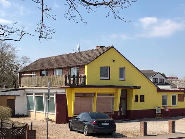 Auf diesem Bild ist eine Immobilie in renovierungsbedürftigen Zustand zu sehen. Das Haus ist im Erdgeschoss mit sehr großen Fenstern ausgestattet im linken Seitenbereich des Hauses befinden sich drei Fenster, im Giebelbereich zwei Fenster und der Hauseingang. Der Bereich mit den großen Fenstern ragt etwas über die Grundmauern des Gebäudes hervor und bildet somit im Obergeschoss einen Balkon an der Längsseite des Hauses und an der Giebelseite dient dieser zur Überdachung des Eingangsbereichs. Die Immobilie wurde auf der rechten Seite mit einem flachen Anbau mit zwei Fenstern versehen. Das Dachgeschoss ist ausgebaut. Die Dachfläche ist asymmetrisch ausgeführt, so dass auf der linken Dachseite zum Balkon hin Fenster und Balkontüren eingebaut sind. Die rechte Dachfläche ist bis zum Anbau im Erdgeschoss heruntergezogen. Die Fassade ist in sonnengelb gestrichen. Der Sockel in dunkelbraun. Die Fassade im Bereich Balkon ist verklinkert. Die Balkonbrüstung ist aus Holz und sehr verwittert. Der Hof vor dem Gebäude ist mit Kieselsteinen belegt. Vor dem Haus parkt ein dunkler PKW.