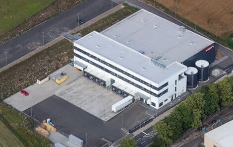 Auf dieser Drohnenaufnahme erkennt man ein sehr großes gewerbliches Gebäude mit Flachdach mit anschließender Halle ebenfalls mit Flachdach ausgestattet, welches als Logistikzentrum genutzt wird. Vor dem Gebäude befinden sich große asphaltierte und betonierte Flächen. Hier können LKW's direkt an die vorgesehenen acht Laderampen des Gebäudes andocken. Über diesen Laderampen befindet sich ein zweigeschossiger Verwaltungstrakt. Der Verwaltungstrakt ist über die komplette Breite des Gebäudes mit Fensterbändern in anthrazit ausgestattet. Die restliche Fassade ist weiß gestrichen. Rechts neben dem Gebäude befinden sich zwei große runde Wasserspeicher ebenfalls anthrazitfarben. Auch die Halle ist anthrazitfarben gestrichen. Rund um das Logistikzentrum sind landwirtschaftliche Flächen zu erkennen. Im Vordergrund sind Bäume abgebildet und die Einfahrt auf das Gelände.