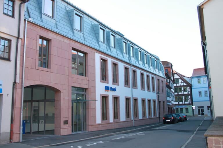 Auf dem Foto ist eine schmale Straße mit Gehwegen und dichter Bebauung teilweise mit Fachwerk zu erkennen. Dominiert wird das Bild durch das neue Gebäude der VR-Bank. Hier handelt sich sich um einen Lückenschluss zwischen zwei Gebäuden. Das Gebäude der VR Bank hat zwei Vollgeschosse und ein ausgebautes Dachgeschoss mit Gauben und moderner Dacheindeckung. Die Fassade ist überwiegend mit Sandsteinen verkleidet ebenfalls wurden die Fensterlaibungen mit Sandstein ausgeführt. Der Rest der Fassade wurde verputzt und weiß gestrichen. Links neben dem Eingangsbereich im Erdgeschoss befindet sich eine Durchfahrt zum Hinterhof.