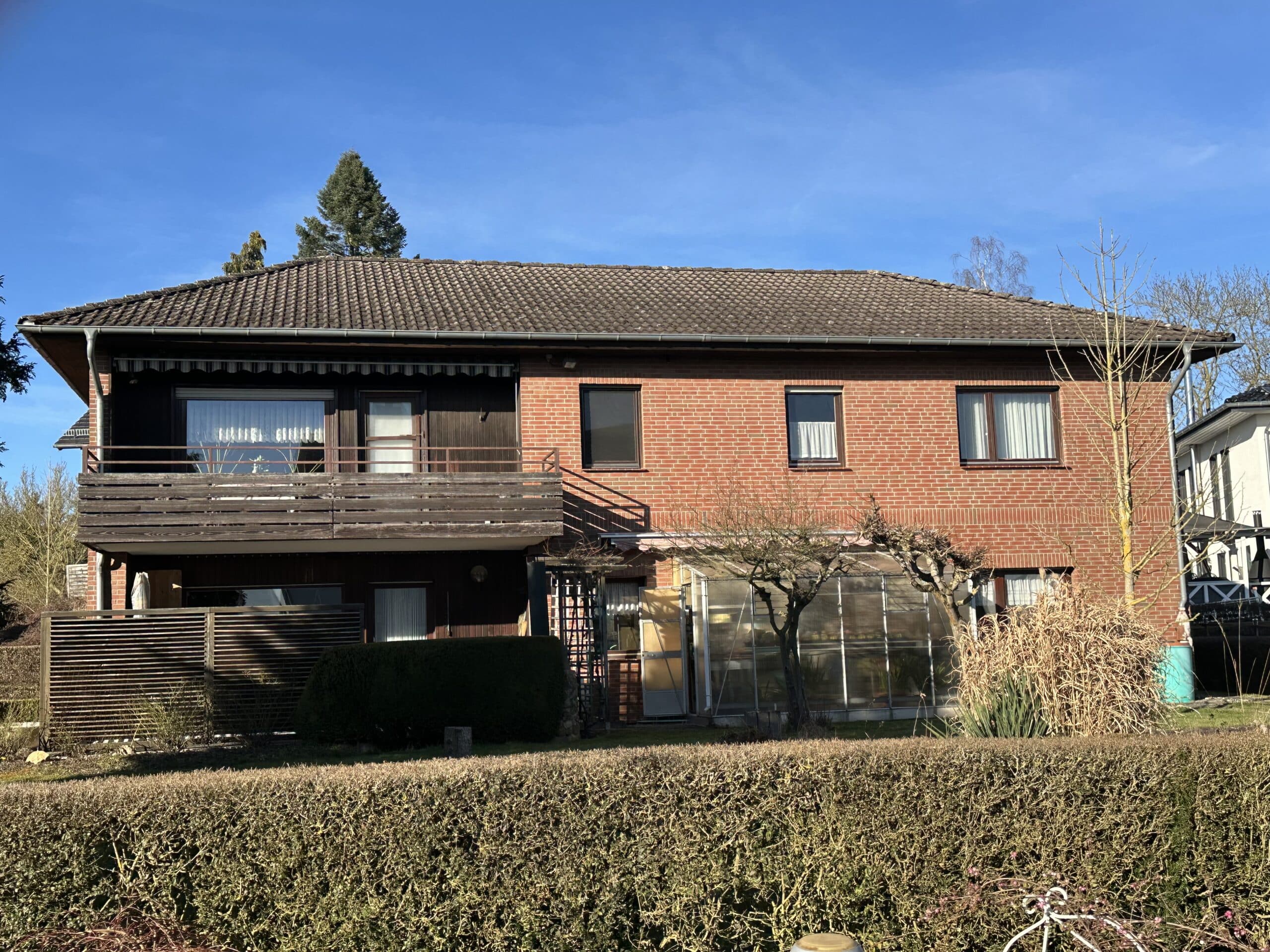 Auf diesem Foto ist ein zweigeschossiges voll verklinkertes Wohnhaus mit Walmdach zu sehen. Im Erdgeschoss der Immobilie befindet sich links eine uneinsehbare Terrasse, darüber ein Balkon. Das Balkongeländer besteht aus horizontalen Holzplanken. Die Außenwand im Balkonbereich ist ebenfalls mit Holz ausgekleidet. Rechts neben der Terrasse befindet sich angelehnt an die Außenwand ein kleineres Gewächshaus. Im Obergeschoss befinden sich baugleich mit dem Unterschoss vier Fenster und eine Balkontür. Im Vordergrund des Hauses ist eine Hecke zu sehen. Rechts im Bild ein Baum. Der Himmel ist blau ohne Wolken.