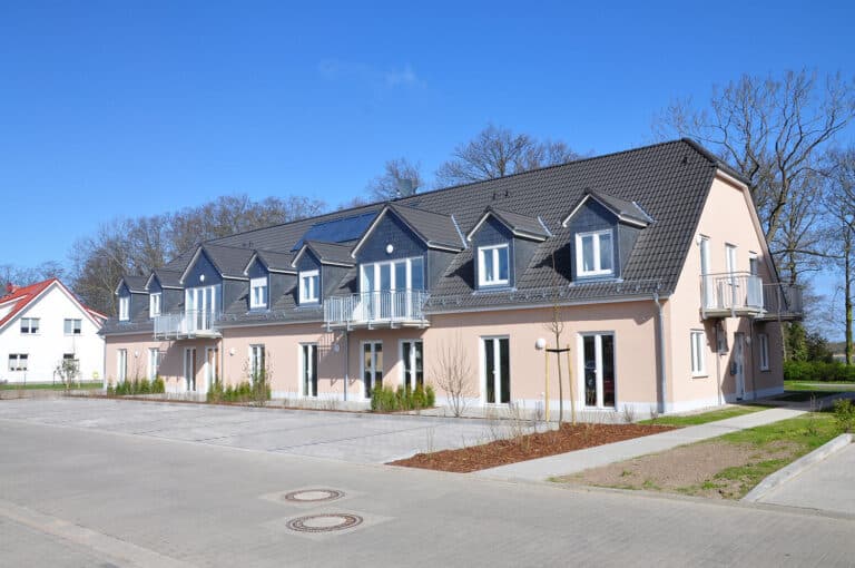 Auf dem Foto ist ein modernes eingeschossiges Wohngebäude mit ausgebautem Dachgeschoss zu sehen. Im Erdgeschoss befinden sich doppelflüglige Terrassentüren. Im Obergeschoss befinden sich sechs Gauben mit Fenstern und zwei größere Gauben mit Ausgang auf einen Balkon. Die Fassade ist in einem Apricot-Ton gestrichen. Die Fenster sind weiß. Das Dach ist mit dunklen Ziegeln eingedeckt. Im Vordergrund befinden sich Parkplätze, sowie teilweise Grünstreifen und Hecken. Die Fußwege, die Parkplätze und auch die Straße vor den Parkplätzen sind gepflastert. Der Himmel ist strahlend blau, ohne Wolken. Im Hintergrund der Immobilie sind kahle Bäume zu sehen.