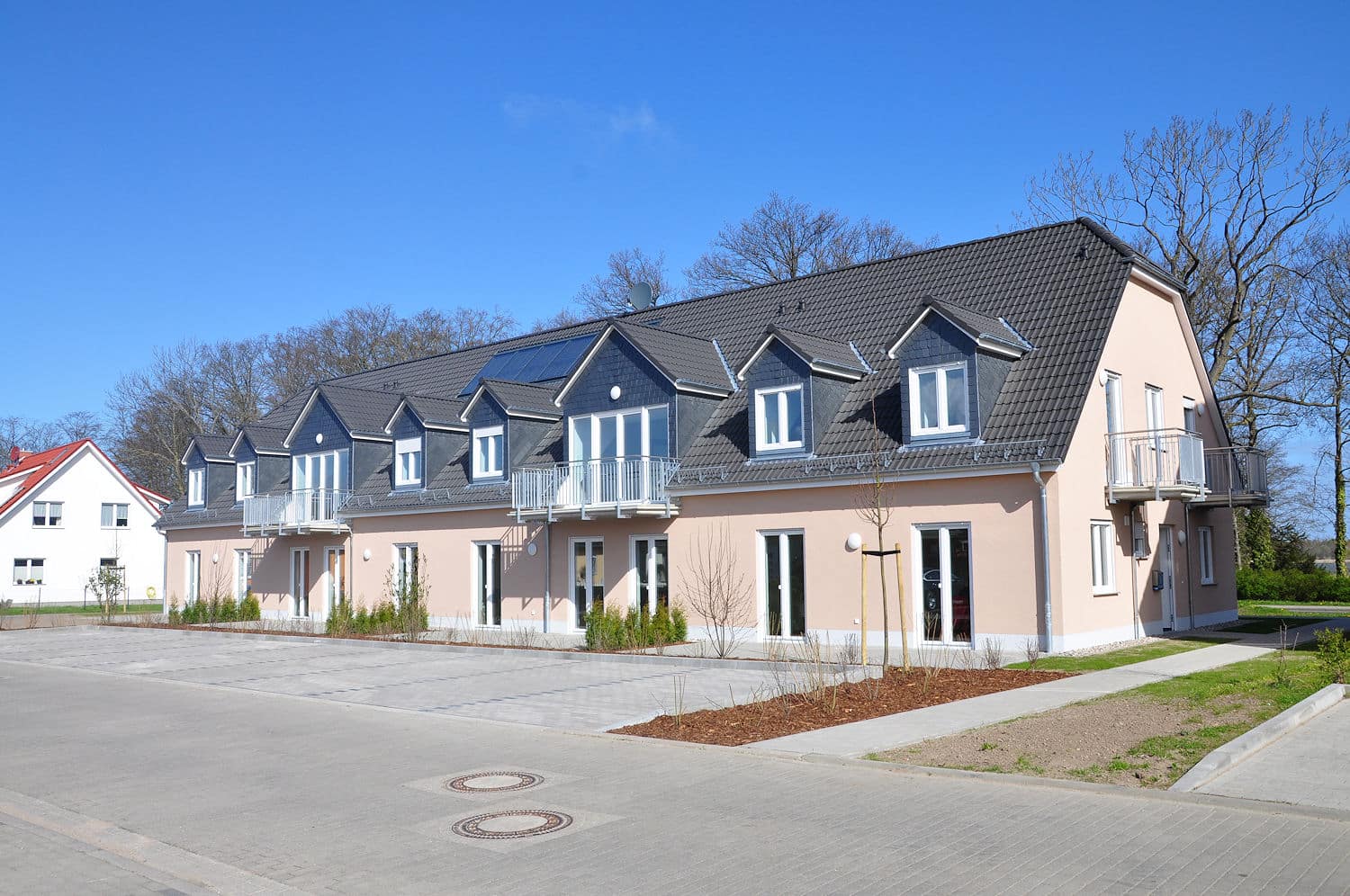 Auf dem Foto ist ein modernes eingeschossiges Wohngebäude mit ausgebautem Dachgeschoss zu sehen. Im Erdgeschoss befinden sich doppelflüglige Terrassentüren. Im Obergeschoss befinden sich sechs Gauben mit Fenstern und zwei größere Gauben mit Ausgang auf einen Balkon. Die Fassade ist in einem Apricot-Ton gestrichen. Die Fenster sind weiß. Das Dach ist mit dunklen Ziegeln eingedeckt. Im Vordergrund befinden sich Parkplätze, sowie teilweise Grünstreifen und Hecken. Die Fußwege, die Parkplätze und auch die Straße vor den Parkplätzen sind gepflastert. Der Himmel ist strahlend blau, ohne Wolken. Im Hintergrund der Immobilie sind kahle Bäume zu sehen.