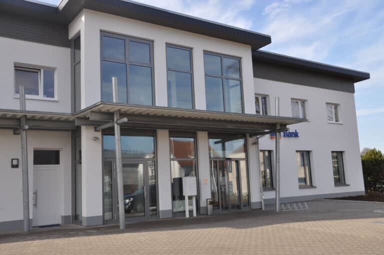 Auf diesem Bild ist ein zweigeschossiges modernes Gebäude mit blauem Himmel und einigen Schleierwolken zu sehen. Das Gebäude hat eine weiße Fassade und steingraue bodentiefe Fenster im großzügigen Vorbau welcher sich in der Mitte des Gebäudes über beide Etagen erstreckt. Über diesen Vorbau gelangt man durch zwei Glastüren die jeweils rechts und links eingebaut sind in das Gebäude. Zwischen den Türen befindet sich ein feststehendes Glaselement und davor eine weiße Briefkastenanlage sowie ein Papierkorb und rechts in der Wand eine Sprechanlage. Im Erdgeschoss befindet sich rechts eine Geschäftsstelle der VR-Bank mit ebenfalls großen steingrauen Fenstern; über diesen Fenstern erkennt man das Logo der VR-Bank. Die Fenster im Obergeschoss sind weiß ausgeführt. Über den Fenstern im Obergeschoss befindet sich eine in dunkelgrau gestrichene circa 80 cm hohe Holzverkleidung. Auf der linken Seite des Haupteingangs befindet sich noch ein Nebeneingang mit Glasoberlicht ansonsten ist die Tür weiß. Der Nebeneingang sowie der Haupteingang werden durch ein Wellblech-Vordach welches auf drei Stahl Rundstützen befestigt ist überdacht. Vor dem Gebäude erkennt man eine gepflasterte Fläche mit Fahrradständer.