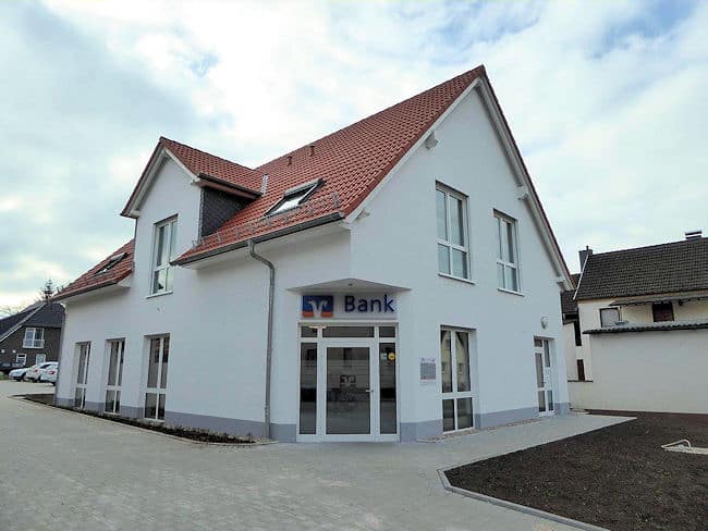 Auf dem Bild ist ein Gebäude zu sehen welches einen Einfamilienhaus entspricht. Dieses Gebäude wird allerdings als Geschäftsstelle der VR-Bank genutzt. Das Erdgeschoss verfügt über bodentiefe Fenster. Über eine abgeschrägte Hausecke führt eine Glaseingangstür in das Gebäude. Über der Tür erkennt man das Logo der VR-Bank. Auch im ausgebauten Dachgeschoss befinden sich bodentiefe Fenster, eine große Gaube und zwei Dachflächenfenster. Das Dach ist mit roten Dachziegeln eingedeckt. Das Gebäude ist weiß gestrichen und im Sockelbereich steingrau. Das Gebäude ist umschlossen von Pflasterflächen. Im rechten Bildrand ist angrenzend an die Pflasterfläche eine ebene Erdfläche ohne Bepflanzung zu erkennen mit angrenzender Mauer. Im linken Hintergrund des Gebäudes erkennt man Parkflächen mit Fahrzeugen sowie ein weiteres Wohngebäude.