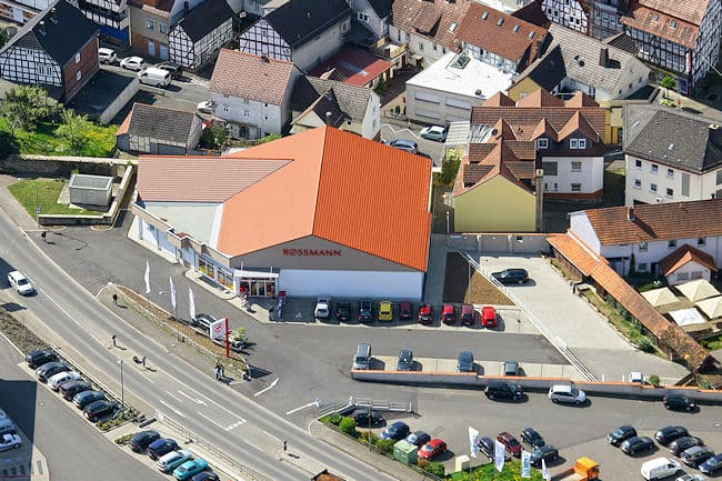 Bei diesem Luftbild erkennt man in der Mitte des Bildes einen Rossmann Markt mit roter Dacheindeckung. Im Vordergrund des Gebäudes befinden sich Parkplätze für den Rossmann Markt und die angrenzenden Gewerbeunternehmen, sowie eine Straße mit Abbiegerspur zum Rossmann Markt. Ebenfalls ist vor dem Gebäude eine Werbeanlage von Rossmann mit drei Fahnen zu sehen. Links neben dem Markt befindet sich hinter einer Mauer eine Trafostation. Rechts neben dem Markt sind ebenfalls Parkplätze für Rossmann eingebaut. Im Hintergrund des Marktes sind Teile der Altstadt mit Fachwerkhäusern zu sehen.