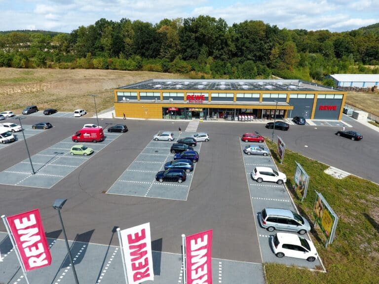 Auf dieser Drohnenaufnahme ist mittig ein Lebensmittelmarkt abgebildet. Im Vordergrund des Marktes ist eine sehr große asphaltierte Parkplatzfläche mit drei Rewe Fahnen zu sehen. Im rechten Bildrand befindet sich die Zufahrt zum Markt und zur Parkplatzanlage. Zwischen der Zufahrt und dem Parkplatz erkennt man eine Grünfläche mit drei großen Werbeschildern. Im Hintergrund und links neben dem Markt befinden sich Felder, Wiesen und Wald. Das Gebäude ist rechteckig mit Flachdach ausgestattet. Die Fassade wurde teilweise mit Trespa-Platten in heller Holzoptik verkleidet beziehungsweise sind Teile der Fassade verputzt und anthrazitfarben gestrichen. Der Haupteingang des Marktes ist überdacht. Hier befinden sich auch die Einkaufswagen. Über der gesamten Breite des Marktes befinden sich Oberlichter. Der Himmel ist blau und mit Wölkchen durchzogen.