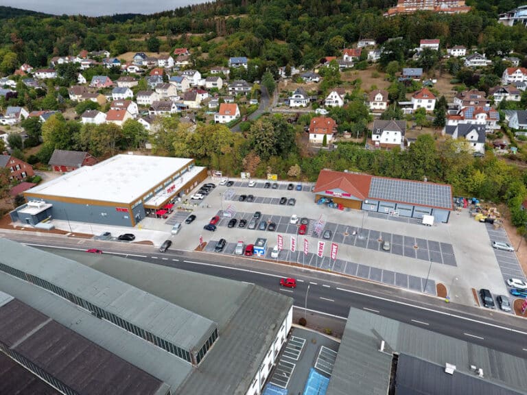 Auf der Drohnenaufnahme ist in der Mitte ein moderner Rewe Markt mit großer Parkplatzanlage und Getränkemarkt zu sehen. Vor dem Gelände verläuft eine Straße. Gegenüberliegend auf der anderen Seite der Straße befinden sich große Hallen und Gebäude. Hinter dem Rewe Markt erkennt man eine Hangbebauung mit überwiegend Einfamilienhäusern und Grünanlagen. Im oberen Bereich des Hanges ist eine sehr große Immobilie zu sehen welche als Hotel oder Klinikum genutzt werden könnte.