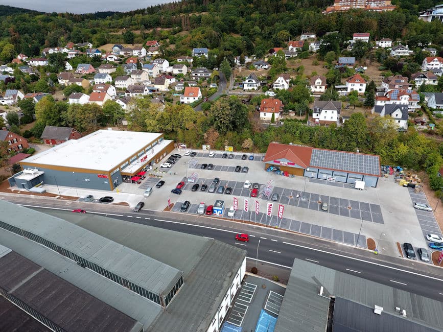 Auf der Drohnenaufnahme ist in der Mitte ein moderner Rewe Markt mit großer Parkplatzanlage und Getränkemarkt zu sehen. Vor dem Gelände verläuft eine Straße. Gegenüberliegend auf der anderen Seite der Straße befinden sich große Hallen und Gebäude. Hinter dem Rewe Markt erkennt man eine Hangbebauung mit überwiegend Einfamilienhäusern und Grünanlagen. Im oberen Bereich des Hanges ist eine sehr große Immobilie zu sehen welche als Hotel oder Klinikum genutzt werden könnte.