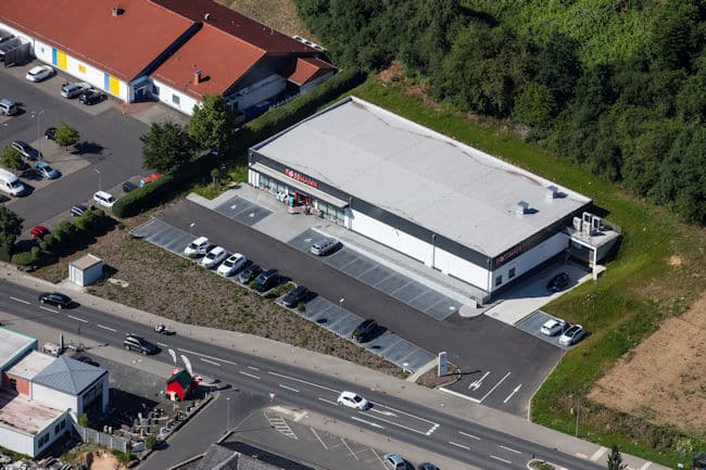 Auf diesem Luftbild ist in der Bildmitte ein großer Drogeriemarkt mit einer Parkplatzanlage zu sehen. Vor der Parkplatzanlage verläuft eine Straße mit Abbiegerspur zum Markt. Auf der gegenüberliegenden Seite des Drogeriemarktes befinden sich weitere Parkplätze und Gewerbegebäude. Im linken Bildrand erkennt man einen Lebensmittelmarkt mit Parkplatz. Hinter und rechts neben dem Drogeriemarkt befindet sich eine Grünfläche. Das Gebäude ist rechteckig. Rechts am Gebäude befindet sich die Anlieferung für LKW. Links befindet sich der Haupteingang mit Glasflächen. Das Gebäude ist weiß gestrichen und im oberen Bereich anthrazitfarben abgesetzt.