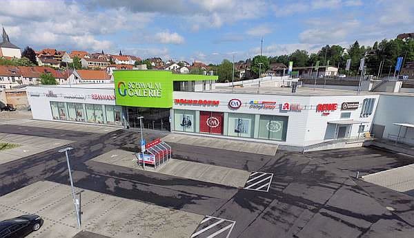 Auf der Drohnenaufnahme ist das Einkaufszentrum Schwalm-Galerie mit der Parkplatzanlage und dem Parkdeck zu sehen. Der Himmel ist blau und mit Wolken durchzogen. Hinter dem Gebäude erkennt man die Stadtsilhouette von Treysa. Das rechteckige Gebäude ist mit Logos im oberen Gebäudeteil bestückt: Rossmann, Vockeroth, ernstings family, Polos Nudeln, mister*lady, REWE, Apotheke, HC Parfümerie, Schwälmer Cafè und über dem Eingangsbereich befindet sich etwas hervorgehoben der Schriftzug Schwalm-Galerie auf maigrünem Hintergrund.
