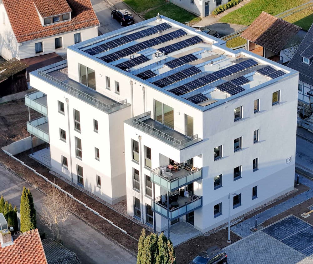 Auf dieser Drohnenaufnahme erkennt man ein viergeschossiges Wohnhaus mit Flachdach und PV-Anlagen. Im Obergeschoss befinden sich Penthouse-Wohnungen mit großzügigen Terrassen. Das Erdgeschoss sowie das 1. und 2. Obergeschoss sind identisch. Alle Wohnungen verfügen über einen Balkon beziehungsweise im Erdgeschoss über eine Terrasse. Die Fassade ist im Bereich Penthouse und Erdgeschoss in einem dezenten Creme-Ton gehalten, das 1. und 2. Obergeschoss in weiß. Links neben dem Gebäude und hinter dem Gebäude erkennt man weitere Wohnhäuser. Das Haus ist sowohl im Norden als auch im Süden von einer Straße umgeben. Östlich der Immobilie befinden sich gepflasterte Parkplätze. Die Außenanlage ist aufgrund der Straßen und Parkplätze sehr beengt.