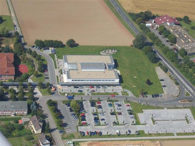 Auf diesem Luftbild ist das Medizinische Versorgungszentrum Treysa mit Parkplatzanlage zu sehen. Das Gebäude wird durch Straßen umringt. Auf der rechten Bildseite erkennt man ein langgezogenes Gebäude welches für eine Werkstatt genutzt wird. Im linken Bereich des Fotos befindet sich ein Klinikgebäude und weitere Mehrfamilienhäuser. Im vorderen Bereich des Gesundheitszentrums befindet sich die große Parkplatzanlage, rechts davon und teilweise auch hinter dem Gebäude erkennt man eine große gepflegte Grünanlage. Hinter dem Grundstück Richtung Westen befindet sich ein großes abgeerntetes Feld.