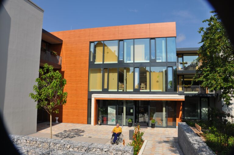 Auf dem Foto ist eine hochmoderne Seniorenwohnanlage mit viel Glasflächen zu sehen. Das Gebäude ist 3-geschossig und mit einem Flachdach ausgestattet. Teile der Fassade sind mit kirschbaumfarbigen Trespa-Platten versehen. Im Eingangsbereich befindet sich ein gepflasterter Vorplatz mit einem Baum und Grünpflanzen, sowie Begrenzungen durch Gabionenkörbe und eine Holzsitzbank. Auf diesem Vorplatz befindet sich ebenfalls ein männlicher Rollstuhlfahrer. Rechts von dem Vorplatz erkennt man einen kleinen Teil einer beginnenden Grünanlage. Links im Bild ist eine graue Hauswand ohne Fenster zu sehen. Der Himmel ist blau.