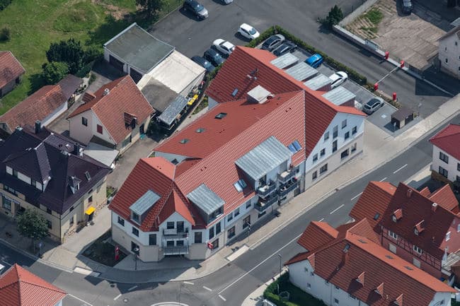 Auf dem Luftbild ist eine Straßenkreuzung mit Kreisel und Markierungen zu sehen, sowie eine umliegende Bebauung mit großen Gebäuden. Die Straße hat rechts und links gepflasterte Gehwege. Die Dächer der Häuser sind überwiegend mit roten Dachziegeln eingedeckt und die mit Gauben ausgestattet. Bei dem zwei- bis dreigeschossigen Gebäuden handelt es sich um Wohn- und Geschäftshäuser mit Gewerbeflächen im Erdgeschoss sowie Wohnungen in den Obergeschossen. In der Mitte des Bildes ist ein sehr großes Eckgebäude mit sechs Balkonen in den Obergeschossen zu sehen. Hinter dem Gebäude befinden sich zehn gepflasterte Parkplätze, sowie auf dem Nachbargelände weitere asphaltierte Parkflächen. Im linken Bildrand erkennt man Grünflächen mit Büschen und Hecken.