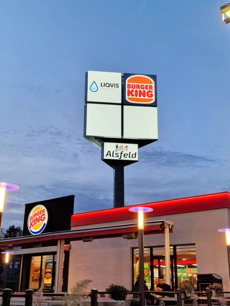 Auf diesem Foto sieht man einen beleuchteten dreiseitigen Werbepylon mit jeweils vier Werbeflächen auf einer Seite. Nur die beiden oberen Werbeflächen sind belegt mit den Logos LIQVIS - oben links und Burger King oben rechts. Die unteren beiden Werbeflächen sind frei. Direkt unter der großen Werbefläche befindet sich noch ein kleines Logo der Stadt Alsfeld. Der Himmel ist dunkelblau mit kleinen Schleierwolken. Im Vordergrund des Pylons erkennt man das Fast Food Restaurant Burger King mit großer beleuchteter Werbeanlage über dem Eingang, sowie drei leuchtende Straßenlampen und eine Terrasse mit Personen.
