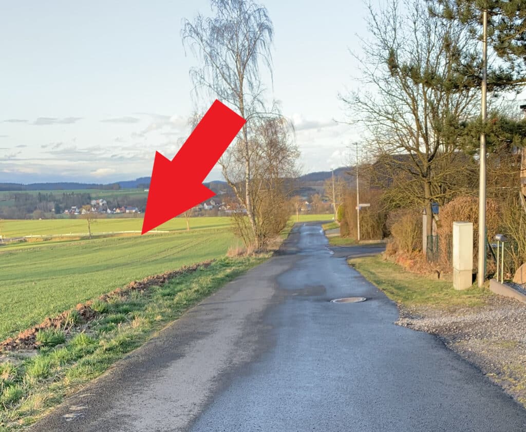 Auf dem Foto ist eine gerade verlaufende asphaltierte Nebenstraße ohne Straßenmarkierungen zu sehen. Die Straße ist stellenweise nass. Rechts neben der Straße befindet sich ein Grünstreifen. Dahinter erkennt man Hecken beziehungsweise eine kleine Mauer und Zäune zu angrenzenden Grundstücken. Ebenfalls befinden sich rechts von der Straße drei Straßenlampen und zwei abgehende Nebenstraßen. Links von der Straße befindet sich ein Graben und anschließend eine gemähte Wiese. Der linke Straßenrand wird von einigen Birken gesäumt. Im weiteren Bildhintergrund ist ein Ort zu erkennen. Dominiert wird das Bild durch einen großen roten Pfeil, welcher auf die Wiese zeigt. Der Himmel ist bedeckt.