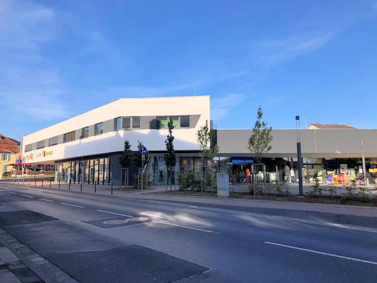 Auf diesem Bild ist ein modernes teilweise zweigeschossiges Gebäude direkt an einer Straße mit Ampelanlage zu sehen. Im Erdgeschoss befinden sich große Schaufenster. Alle Räumlichkeiten sind vermietet und die Schaufenster üppig dekoriert. Das Obergeschoss kragt etwas über das Untergeschoss. Hier befinden sich weitere Geschäftsräume. Die Fassade im Obergeschoss ist weiß mit grauen Akzenten im Fensterbereich gestrichen. Die Fenster sind grau und bei drei Fenstern sind die Jalousien geschlossen. Das Obergeschoss ist nur zu einem Drittel ausgebaut. Der nicht bebaute Bereich im Obergeschoss dient als Parkdeck. Zwischen dem Gebäude und der Straße befindet sich ein Gehweg und ein Grünstreifen mit Bäumen. Der Himmel ist blau. Die Straße liegt im Schatten.