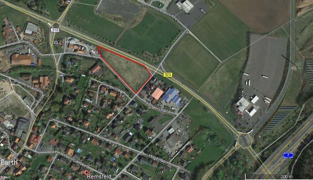 Bei diesem Foto handelt es sich um eine Google-Earth Teilaufnahme von Knüllwald-Remsfeld. Am rechten unteren Bild ist ein kleines Teilstück der Autobahn A7 zu erkennen. Parallel dazu befindet sich ein Verkehrskreisel. Von diesem Kreisel ausgehend gelangt man auf die Bundesstraße 323. Im weiteren Verlauf dieser Bundesstraße Richtung Norden befindet sich auf der linken Seite der Straße ein fast dreieckiges Grundstück welches rot markiert ist. Hier befindet sich unser Gewerbegrundstück umschlossen von den Straßen Grüner Weg, Lange Straße und natürlich der Bundesstraße 323. Ebenfalls zu erkennen sind auf der Aufnahme die Straßen Brundershausner Weg, Hinter der Gansbett und Hilgenborn. Hier handelt es sich um ein Wohngebiet überwiegend mit Einfamilienhäusern.
