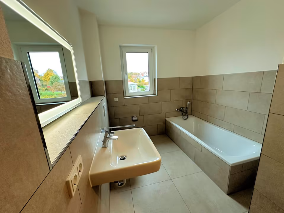 Badezimmer - Schwalm-Terrassen Auf diesem Bild sieht man ein Badezimmer. Das Badezimmer ist halbhoch in einem Beigeton gefliest. Der obere Bereich ist verputzt und weiß gestrichen. Auf der Rechtenseite befindet sich eine Badewanne. An der Kopfseite sieht man die Armatur der Badewanne. Gegenüber der Badewanne befindet sich ein WC und ein Waschbecken. Über dem Waschbecken ist ein beleuchteter Spiegel angebracht. Die Beleuchtung ist eingeschaltet. Links neben dem Waschbecken befindet sich eine Doppelsteckdose. Es handelt sich um ein Tageslichtbad. Das Fenster ist auf der Stirnseite des Raumes.