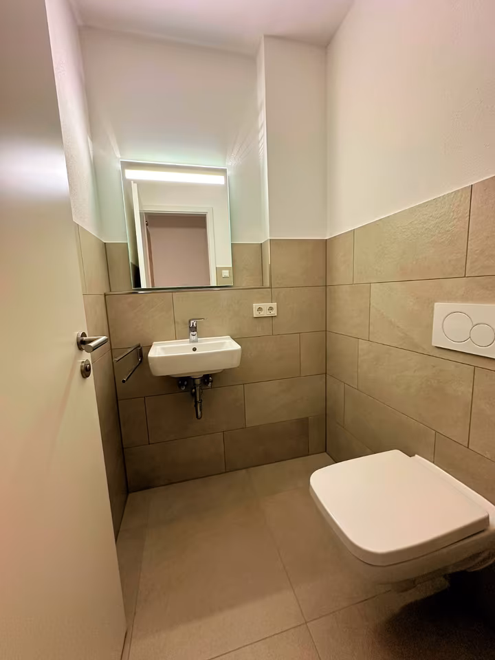 Gäste-WC Schwalm-Terrassen Auf diesem Bild ist ein Gäste-WC zusehen. Die Wände sind halbhoch in beige gefliest. Der obere Bereich ist verputzt und weiß gestrichen. An der Kopfseite des Raums ist ein kleines Waschbecken zu sehen, über dem Waschbecken hängt ein beleuchteter Spiegel. Das Licht ist eingeschaltet. Rechts neben dem Waschbecken befindet sich eine Doppelsteckdose und links ein Handtuchhalter. Rechts im Bild ist ein an der Wand hängendes WC zu erkennen darüber befindet sich die WC-Spülung.
