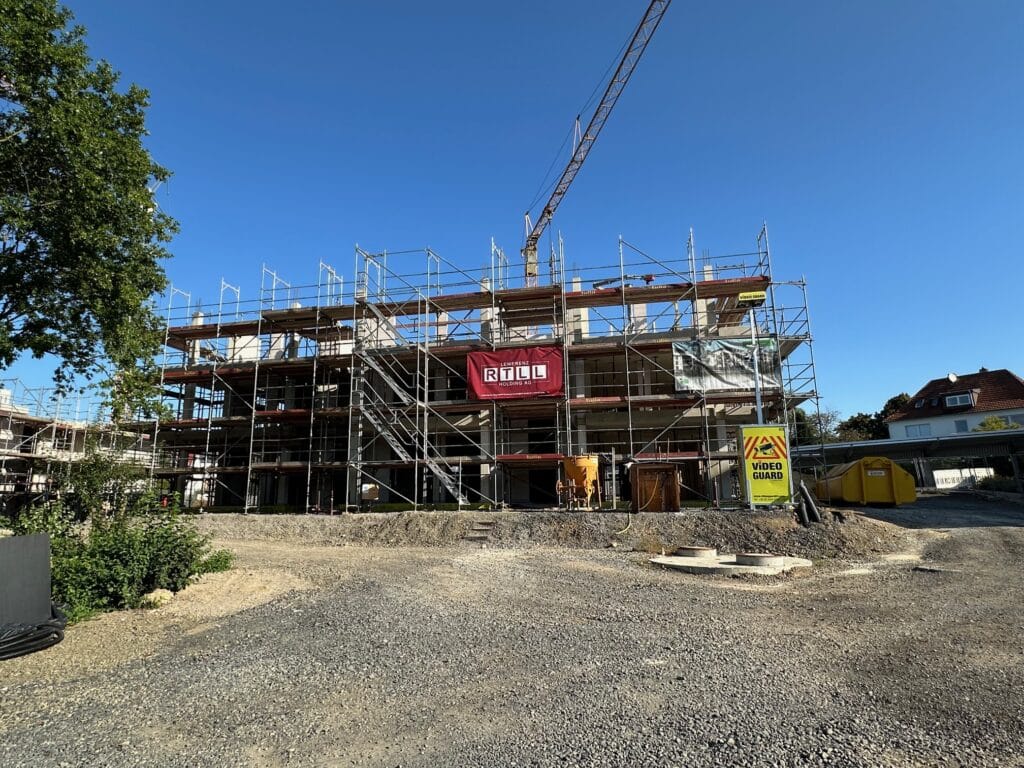 Das Bild zeigt eine Baustelle an einem sonnigen Tag. Im Zentrum des Bildes befindet sich ein Gerüst, das ein noch im Bau befindliches Gebäude umgibt. Das Gerüst ist komplex aufgebaut und besteht aus Metallrohren und -brettern, die den Arbeitern Zugang zu den verschiedenen Stockwerken ermöglichen. Das Gebäude selbst ist noch in der Rohbauphase, mit Betonwänden und -pfeilern, die die Struktur bilden. Auf dem Gerüst sind mehrere Banner angebracht, darunter eines mit dem Logo "RTLL" und dem Zusatz "LEWERENZ HOLDING AG". Ein großer Baukran erstreckt sich vom oberen rechten Bildrand und ragt über das Gebäude hinaus. Er wird verwendet, um Baumaterialien anzuheben und zu positionieren. Der Himmel ist strahlend blau und ohne Wolken. Im Vordergrund befindet sich ein Bereich mit Schotter und Erde, der die Baustelle umgibt. Mehrere Baugeräte und -materialien, wie gelbe Container und ein Hinweisschild mit der Aufschrift "VIDEO GUARD", sind auf der Baustelle verstreut. Links im Bild ist ein Baum mit dichtem grünen Laub zu sehen. Im Hintergrund sind weitere Gebäude zu erkennen, darunter ein Wohnhaus mit rotem Dach.