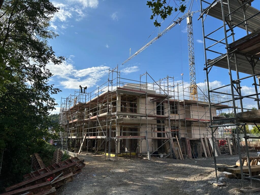Das Bild zeigt eine Baustelle unter einem strahlend blauen Himmel mit einigen weißen Wolken. Im Zentrum des Bildes befindet sich ein Gebäude, das sich im Bau befindet. Es ist von einem Gerüst umgeben, das den Arbeitern den Zugang zu den verschiedenen Stockwerken ermöglicht. Ein großer Kran ragt in den Himmel und dominiert die rechte Seite des Bildes. Er ist gelb und weiß lackiert und wird vermutlich zum Heben schwerer Baumaterialien verwendet. Der Vordergrund des Bildes ist mit Schotter bedeckt, auf dem sich Baumaterialien wie Holzbretter befinden. Auf der linken Seite des Bildes sind Bäume zu sehen, die einen grünen Rahmen um die Szene bilden. Das Gerüst und der Kran sind in hellen Grautönen gehalten, während das Gebäude selbst einen hellen, neutralen Farbton aufweist.