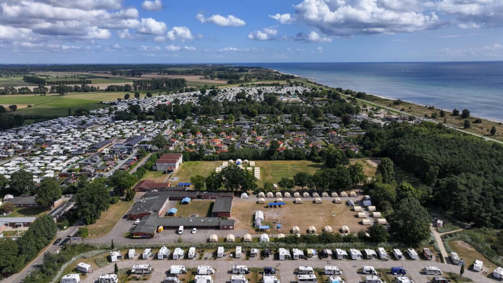 Das Bild zeigt eine Luftaufnahme eines Küstengebietes, das eine harmonische Mischung aus Natur und menschlicher Besiedlung einfängt. Auf der linken Seite des Fotos sehen wir eine weite Fläche üppig grüner Felder. Zwischen diesen Feldern gibt es eine dichte Anordnung weißer Wohnmobile und Wohnwagen, die ordentlich auf ausgewiesenen Flächen geparkt sind, was auf einen Campingplatz oder Ferienpark hinweist. In Richtung Mitte zeigt das Bild einen rasterartigen Grundriss von Häusern mit unterschiedlichen Dachkonstruktionen. Die Farbpalette der Dächer der Häuser umfasst Rot-, Braun-, Schwarz- und Grautöne. Dieser Abschnitt wird von einer großen, offenen Fläche mit trockenem Gras begrenz. Rechts des zentralen Campingplatzes liegt eine bewaldete Zone, die einen natürlichen Kontrast zur bebauten Umgebung bildet. Jenseits der Baumgrenze beobachten wir einen unberührten Küstenabschnitt, der unter einem teilweise bewölkten Himmel auf das ruhige, blaue Meer trifft. Das Meer scheint ruhig zu sein mit leichten Wellen auf der Oberfläche.