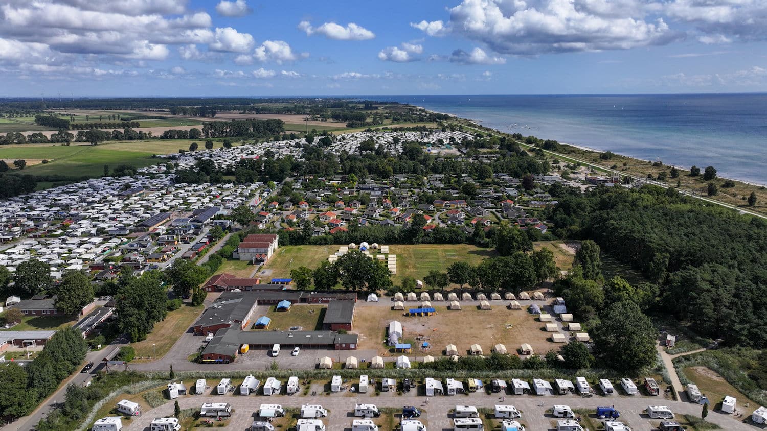 Das Bild zeigt eine Luftaufnahme eines Küstengebietes, das eine harmonische Mischung aus Natur und menschlicher Besiedlung einfängt. Auf der linken Seite des Fotos sehen wir eine weite Fläche üppig grüner Felder. Zwischen diesen Feldern gibt es eine dichte Anordnung weißer Wohnmobile und Wohnwagen, die ordentlich auf ausgewiesenen Flächen geparkt sind, was auf einen Campingplatz oder Ferienpark hinweist. In Richtung Mitte zeigt das Bild einen rasterartigen Grundriss von Häusern mit unterschiedlichen Dachkonstruktionen. Die Farbpalette der Dächer der Häuser umfasst Rot-, Braun-, Schwarz- und Grautöne. Dieser Abschnitt wird von einer großen, offenen Fläche mit trockenem Gras begrenz. Rechts des zentralen Campingplatzes liegt eine bewaldete Zone, die einen natürlichen Kontrast zur bebauten Umgebung bildet. Jenseits der Baumgrenze beobachten wir einen unberührten Küstenabschnitt, der unter einem teilweise bewölkten Himmel auf das ruhige, blaue Meer trifft. Das Meer scheint ruhig zu sein mit leichten Wellen auf der Oberfläche.