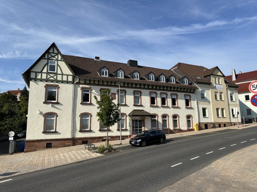 Zu sehen ist ein unter Denkmalschutz stehendes Bürogebäude mit hellem Anstrich und symmetrisch eingebauten Fenstern über die gesamte Front. Im vorderen Bereich des Gebäudes befinden sich vier PKW-Stellplätze, ein Baum, eine Sitzbank und ein Briefkasten. Der Eingangsbereich im Erdgeschoss ist überdacht und von rechts über eine Rampe und von links mit zwei Stufen zu erreichen.